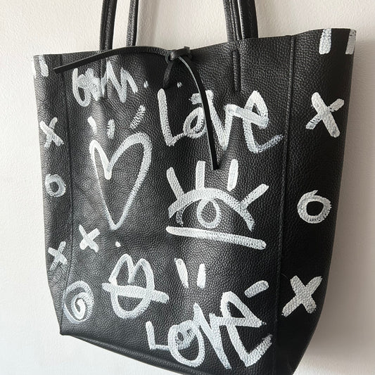 Black leather tote bag with white graffiti-style designs on a white background